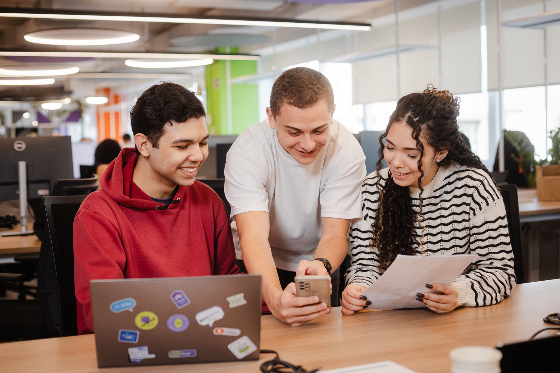 Três pessoas do time do Grupo OLX olhando juntas uma folha e um celular, com um notebook cheio de adesivos na mesa