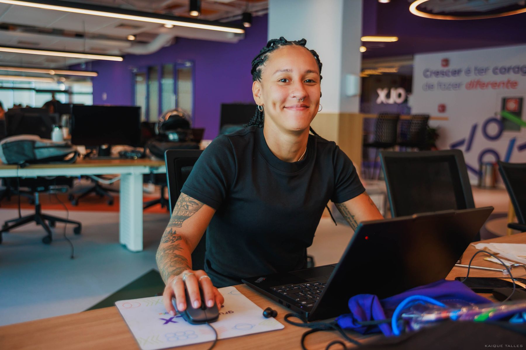 Mulher de camiseta preta sorrindo, com a mão no mouse, diante de um notebook